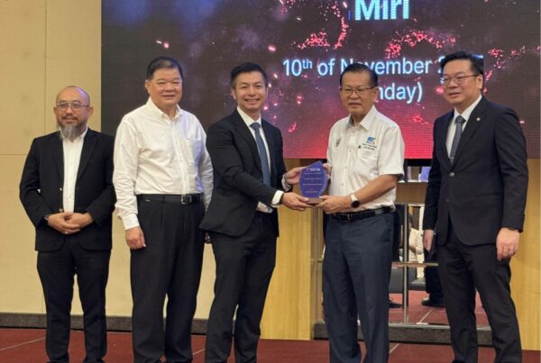 A group of five men in formal attire stand on a stage during an event. The man in the center in a suit holding a plaque from another man wearing a white shirt, while others stand on both sides. A large digital backdrop behind them displays event details.